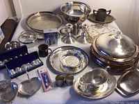 Overview shot of silver plated serving items including trays, bowls, salt and pepper sets, candlesticks, a gravy boat, covered dish, metal cup, and trivet.