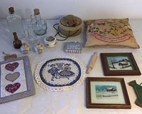 Photo showing a vintage embroidered floral pillow, vintage hat box with floral ribbons, assorted old glass bottles, framed textile pieces, textile hearts, and a small wooden rolling pin and sign on a white surface.