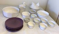 All ceramic dishes arranged on table with purple plates in front and white dishes, ramekins, and lidded figurine dishes behind.