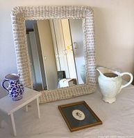 Wicker mirror with floral print frame, two ceramic pitchers, and small stool arranged on top of a surface