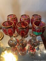 Six matching ruby red overlay crystal wine goblets with gilt decoration displayed on mirrored surface
