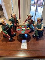 Five clown figurines seated around a round poker table, each holding playing cards.