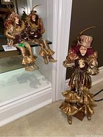 Two seated clown dolls in gold and maroon costumes perched on a glass shelf with mirrored reflection