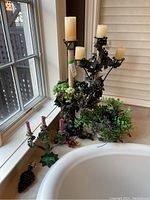 Photo showing large multi-arm candleholder with cream pillar candles, smaller three-arm taper candleholder with red candles, and grapevine decor with green and purple grapes on bathroom counter near window.
