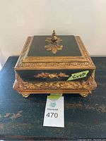 Front and top view of square decorative jewelry box with ornate gold trim and finial, placed on a dark surface with a measuring label.