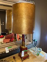 Full lamp with gold shade on top of wooden table, visible mirror and other decorative glass items in background.
