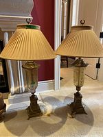 Pair of two decorative table lamps with tall beige pleated shades and ornate metal bases placed on carpet.