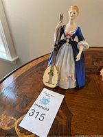 Full front view of the figurine on wooden table showing lady holding lute and auction tag