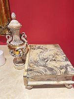 Photo showing vase with sculpted cherubs in white and gold tones beside decorative wooden box with classical artwork.