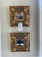 Two square wall mirrors stacked vertically on a wall with ornate gold leaf textured frames, each mirror center reflecting part of room.
