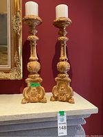 Two carved wooden candle holders with white pillar candles on top, placed on a stone mantel against a dark red wall near a framed artwork.