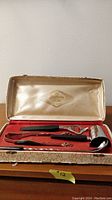 Open box showing four bar utensils with black Bakelite handles resting in red felt interior with satin lid featuring gold logo.
