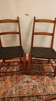 Front view of two antique Shaker-style chairs with black woven seats placed on a patterned rug against a white wall.