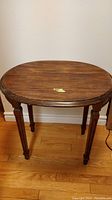 Front view of oval wooden antique table showing top surface and carved legs, placed on wooden floor against wall.