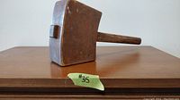 Side view of antique wooden mallet showing rectangular head and straight handle on wooden surface.