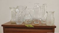 Nine assorted glass and crystal vases of varying heights and designs shown on wood surface.