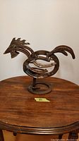 Single view of the handmade metal rooster sculpture standing on a wooden table, showing its full profile, including head, body, tail, and circular base.