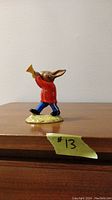 Front view of the Royal Doulton Bunnykins trumpeter figurine showing the rabbit in a red jacket and blue pants, blowing a golden trumpet, standing on a green base.