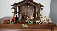 Full view of complete vintage nativity scene set up in front of wooden stable with bark roof. Figures include Mary, Joseph, Baby Jesus, shepherds, animals. Light fixture under roof visible.