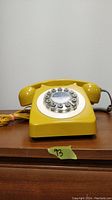 Front side view of mustard yellow rotary phone showing handset, dial, and cord on wooden surface against plain wall.