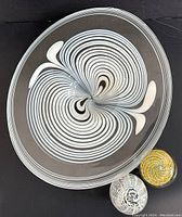Top-down view showing the large platter with white and black swirl design accompanied by two distinct paperweights, one yellow swirled, one black and white patterned.