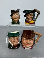 Photo showing four mini Toby jugs placed on a clear stand, showcasing their hand-painted details and facial expressions.