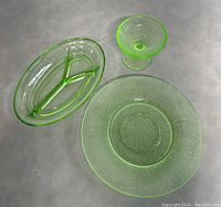 Three pieces of vintage uranium depression glass on a neutral surface: an oval divided dish, a small pedestal compote, and a textured round plate.