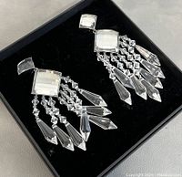 Close-up of one oversized dangly clip-on earring showing faceted square elements and hanging crystal beads against black background box.