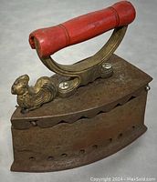 Cast metal charcoal iron with red wooden handle viewed from side showing rooster decoration and ventilation holes.