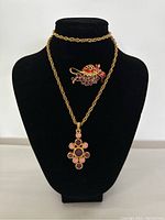 Two images showing a gold-tone ladybug brooch and a gold-tone chain necklace with a floral pink pendant displayed on a black bust stand.