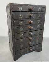 Front view showing seven drawers with round wooden knobs and metal floral decorative accents on each drawer front.