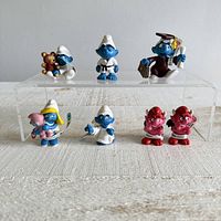 Front view of all 7 Smurf figurines arranged on two clear tiers showing details, colors and expressions.