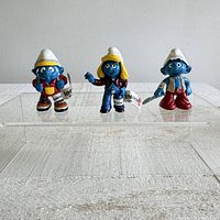 Front view of three Schleich Smurf figurines: Hiker Smurf, Smurfette Trendy, and Techno Smurf, standing on clear acrylic display with visible tags attached.