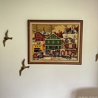 Full view of framed embroidery depicting a town on a wall from a distance.