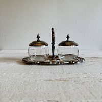 Front view of the silver plated brass condiment caddy with two glass jars, showing decorative tray, central handle and finial lids.