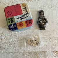 Fossil watch with tin box and extra links accessory package shown on textured surface.