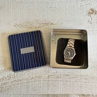 Fossil tin box with watch inside, watch resting on black foam insert, blue striped top cover visible.