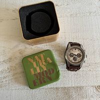 Photo showing Fossil watch with brown leather strap, beige dial, original green metal Fossil box beside it on a light surface.