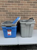 Three Rubbermaid storage bins in blue and gray with lids placed on a table in front of a brick wall.