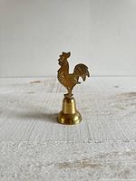Front view of the vintage brass bell with a rooster figure on top, showing the full height and base.