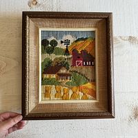 Full framed embroidery artwork held up for display showing entire farm scene with barn, farmhouse, trees, wheat bundles, and windmill.