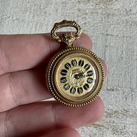 Front view of miniature Hudson gold-toned pocket watch pendant showing dial with Roman numerals and ornate hands held between fingers.