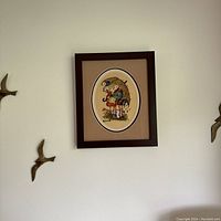 Framed vintage Hummel embroidery mounted on beige textured mat in dark brown frame, shown hanging on wall with bird wall decorations nearby.