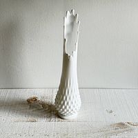 Front view of vintage white milk glass hobnail swung vase on textured surface against plain wall.