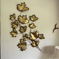 Wall art showing multiple gold-toned metal maple leaves connected by dark stems hanging on a wall with a lamp and bird decoration nearby.