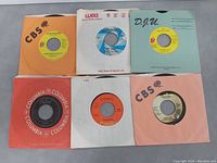 Top view of six 45 RPM vinyl records laid out with sleeves visible, showing labels CBS, WEA, D.Y.U., Columbia, Capitol, CBS. Used vintage condition with minor wear.