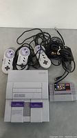 Super Nintendo console with 2 controllers, AV cables, and game cartridge shown together on a flat surface.