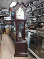 Front view of vintage Tempus Fugit grandfather clock, showing ornate clock face with Roman numerals, wood casing, and glass door revealing pendulum and weights.