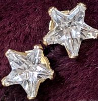 Close-up front view of two star-shaped earrings with clear faceted stones in 9K gold setting on a dark red fabric background.