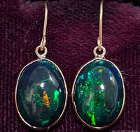 Pair of oval enhanced black opal earrings set in 10K gold with hook fastenings, displayed on a dark purple fabric background.